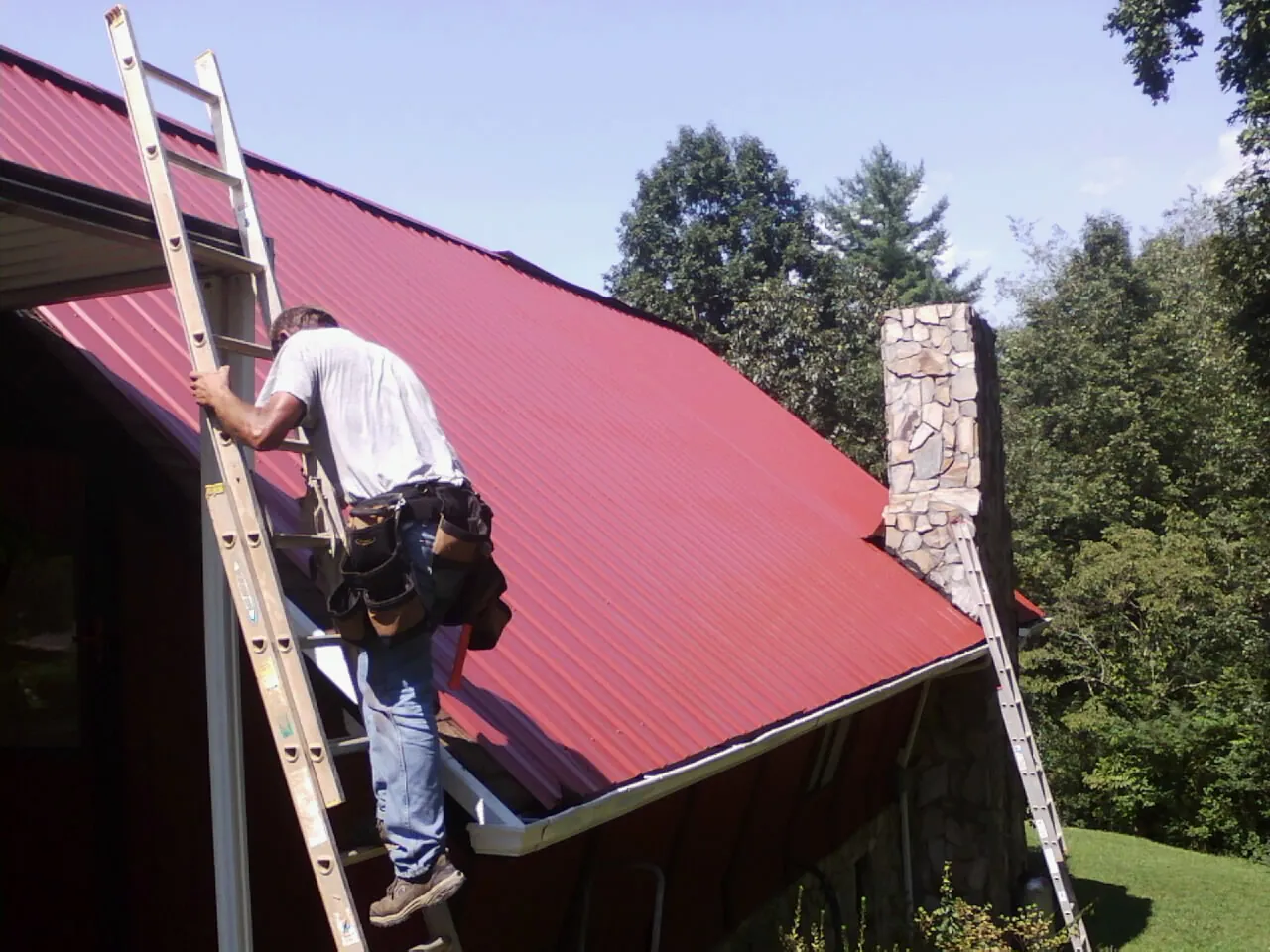 Roofer on a ladder beside a red metal roof and stone chimney — Soffit & Fascia Repair by Roofing Brothers in Plattsburg